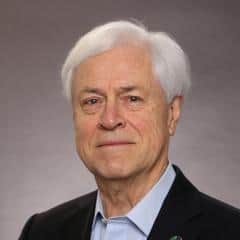 A portrait of Herman Mulder, an elderly white man, standing in front of a neutral light grey background. He is dressed in a black blazer and a light blue shirt, and is wearing a pin on his left lapel representing the United Nations' Sustainable Development Goals. His expression is neutral